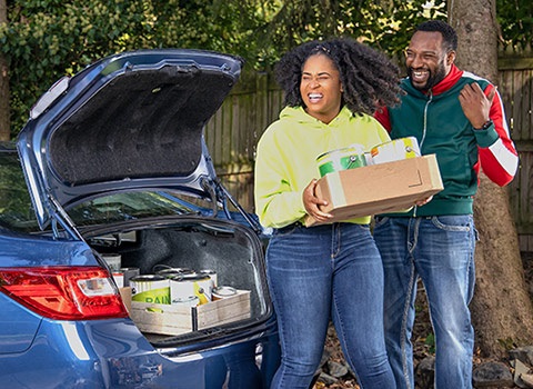 Black woman holding box of paint to recycle, next to Black man and car with trunk open, containing more cans of paint