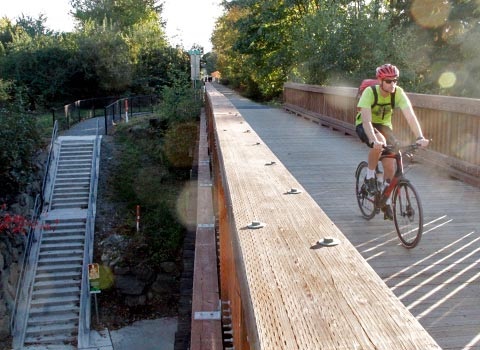 male caucasian cyclist in yellow shirt with red helmet and sunglasses crossing bridge on Cross Kirkland Corridor with stairs down to street visible beside in Houghton neighborhood