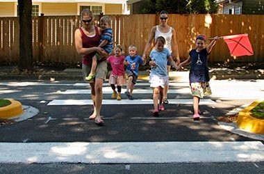families with moms and children crossing the street at a crosswalk with a grassy median, with a little girl holding a pedestrian flag